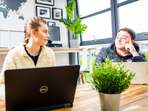 Two business manangement students sitting at a table with their laptops talking about a project