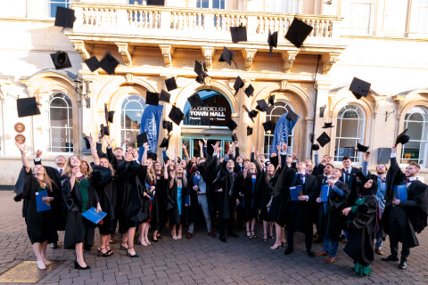 Group of graduating students outside Loughborough Town Hall dressing in graduation gowns throwing their morterboard hats in the air in celebration