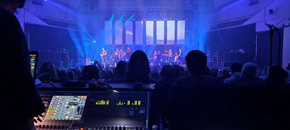 A view of a band on stage from a sound board at the back of a crowd