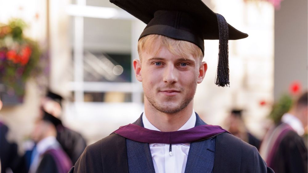 Ethan, a young man with blonde hair looks at the camera. He is wearing a mortarboard cap and his graduation gown. His shirt is white