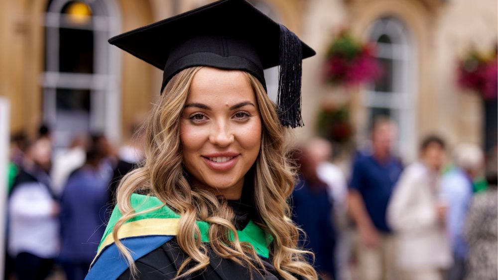 Georgia, a young woman with curled, light brown hair smiles at the camera. She is wearing her mortarboard cap and graduation gown. Her sash colours are blue, yellow and green. 