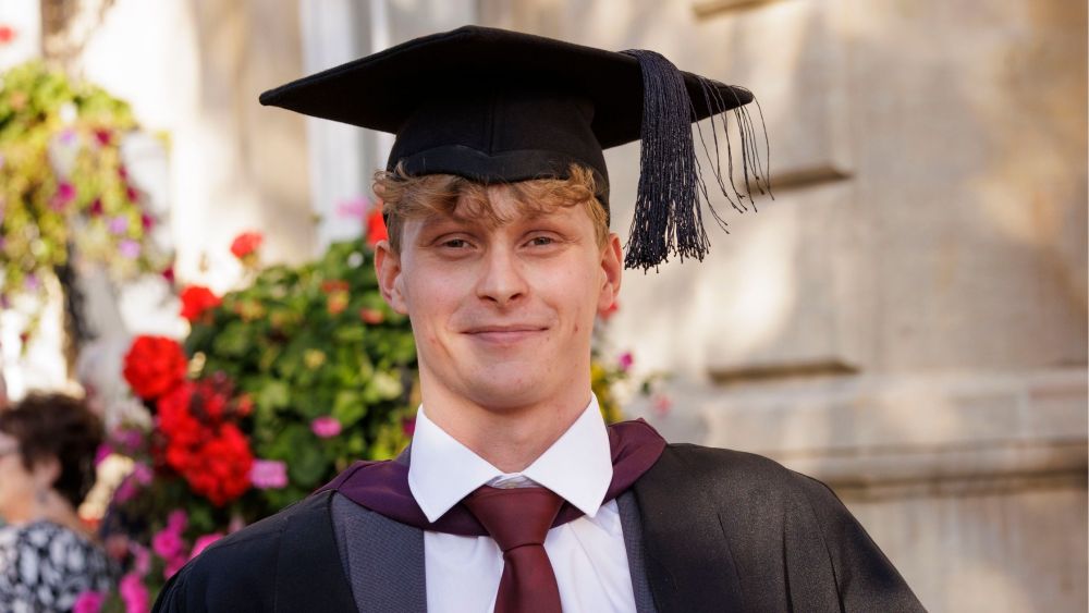 Oliver Smith smiles at the camera, he is wearing a mortarboard cap and graduation gown. He has a red tie and white shirt