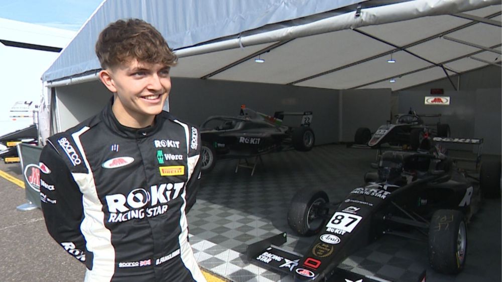 Deagen stands smiling in front of a tent on the left hand side of the image. He is wearing his racing gear. In the tent behind him is his racecar. 